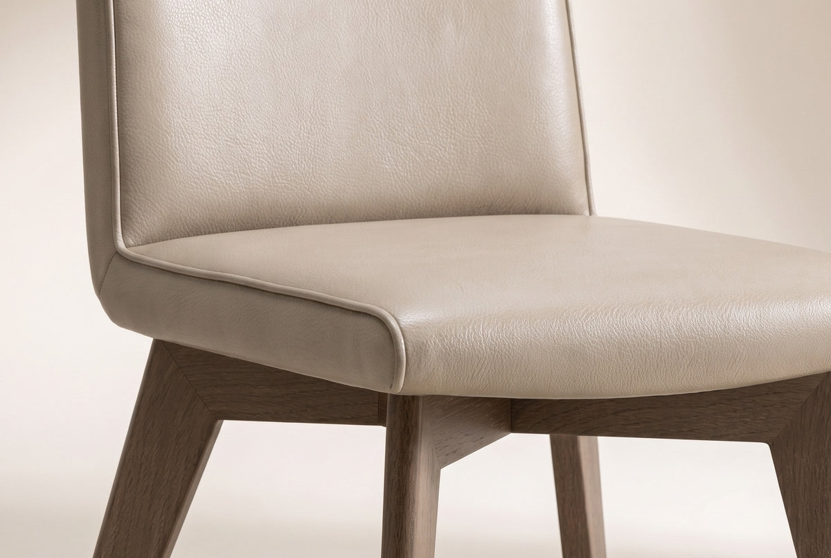 Close-up of one beige Aileen leather dining chair on an off-white backdrop: the pebble-grain leather back and seat, the self-colour piping along the edges, the ash underframe and the tops of the splayed legs