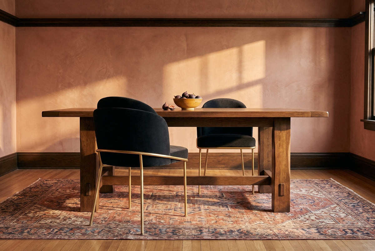 Alexander black velvet dining chair with brass legs seen from behind at a dark oak table with a bowl of figs, a second chair across, terracotta plaster walls and an antique rug in golden-hour light