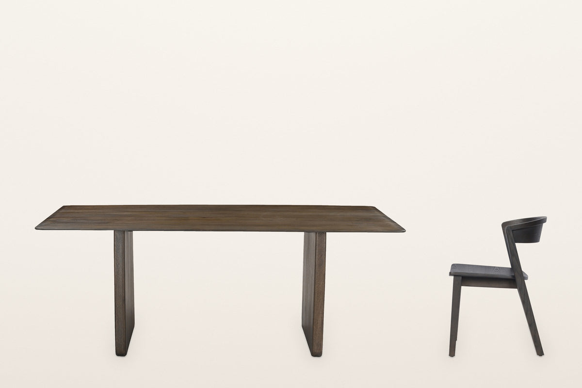 The Arquata table along its long side beside one Certosa chair in profile: a thin top on two slab legs, the chair's back band level with the top