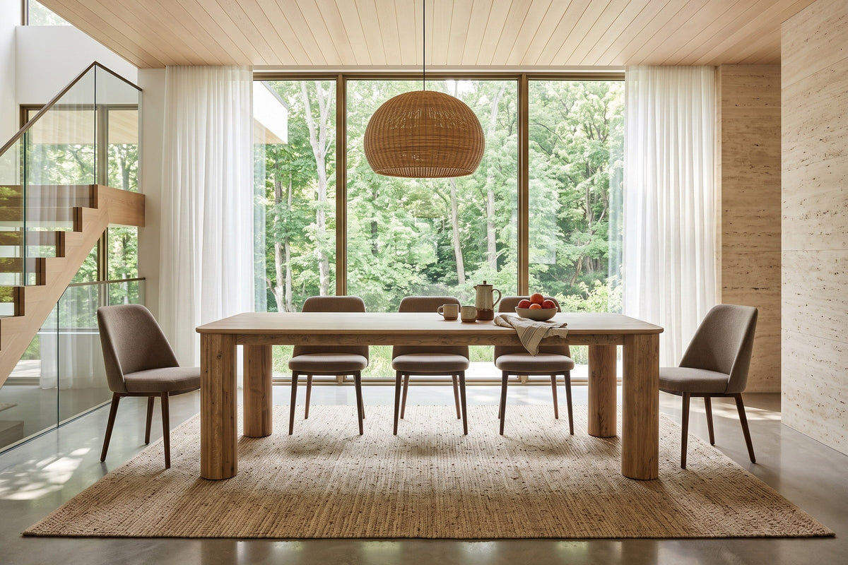 Atzara dining table in a Caledon Hills house, with a tall glass wall and sheer curtains to a maple forest, a travertine wall