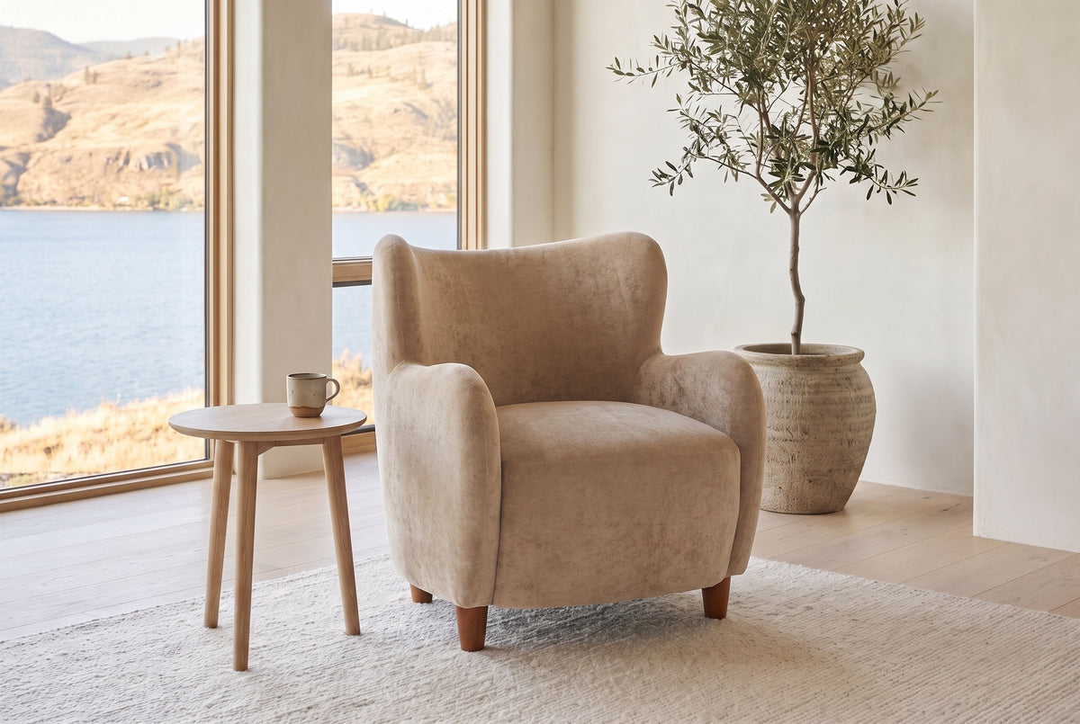 Bormio chair in light camel velvet beside a pale oak side table in a bright lake-house living room with an olive tree