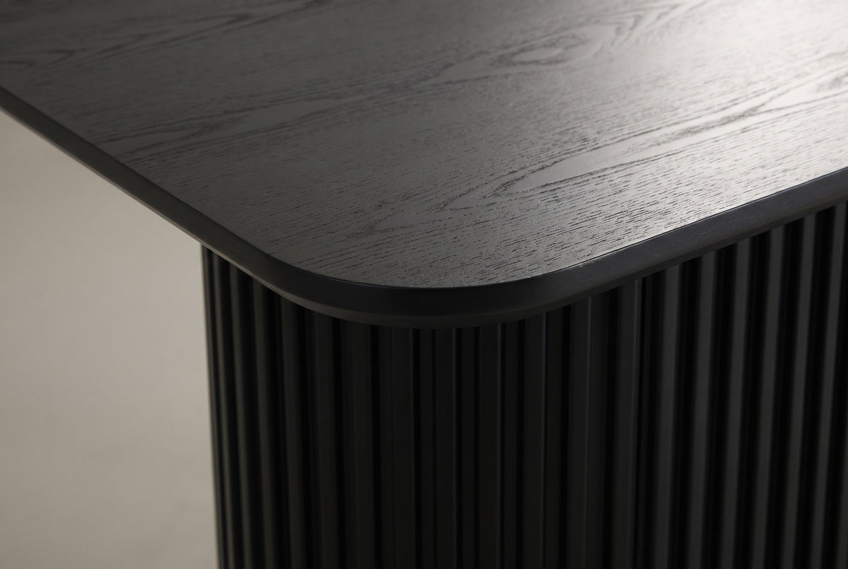 Close-up of the black open-grain top: a softly rounded corner, a slim edge and the fluted pedestal below