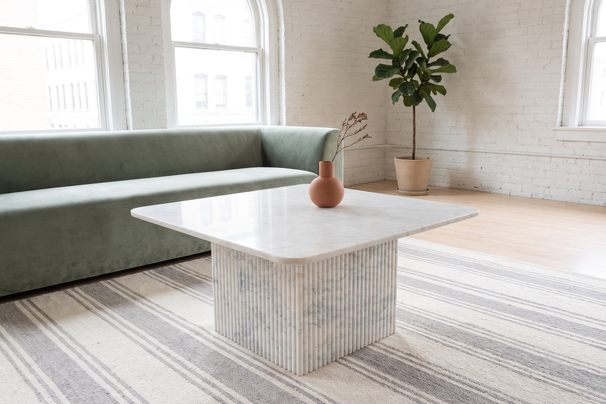 Campiglia white marble coffee table on a striped rug in front of a sage velvet sofa in a white-brick Winnipeg loft with arche