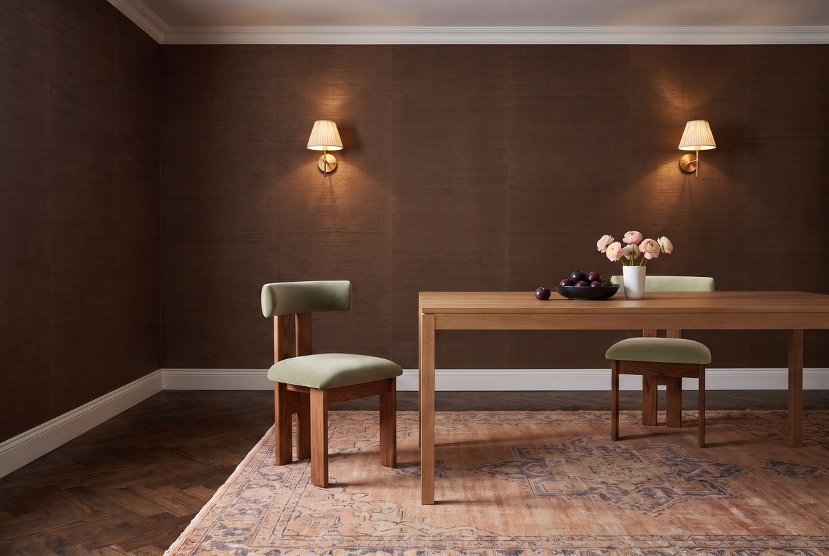 Two Carlo oak dining chairs with sage-olive velvet bolster backs and seats at a pale wood table in an evening dining room with brown grasscloth walls, two lit wall sconces, a faded rug, a bowl of plums and pink ranunculus