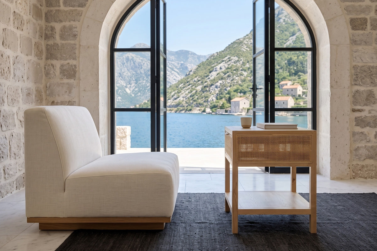 Side view of the Cetara side table in Natural ash and wicker beside an ivory lounge chair in a stone house by the bay
