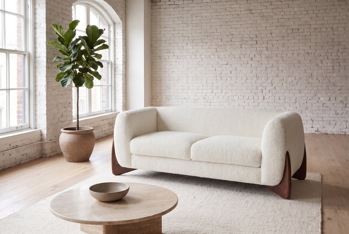 Greccio boucle loveseat in beige in a white-brick loft with arched windows, a fig tree and a round travertine table