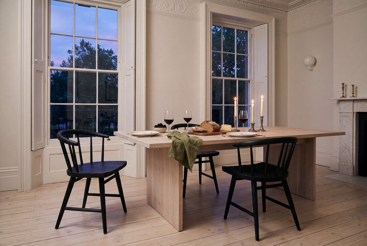 Three black Lauryn dining chairs at a pale oak slab-leg table with lit candles in pewter candlesticks, red wine, bread, cheese, blackberries and a green linen napkin, by tall sash windows with folding shutters, a white marble fireplace with brass candlesticks and an ornate plaster cornice, on wide pale floorboards, in a Georgian townhouse in Dublin in the evening