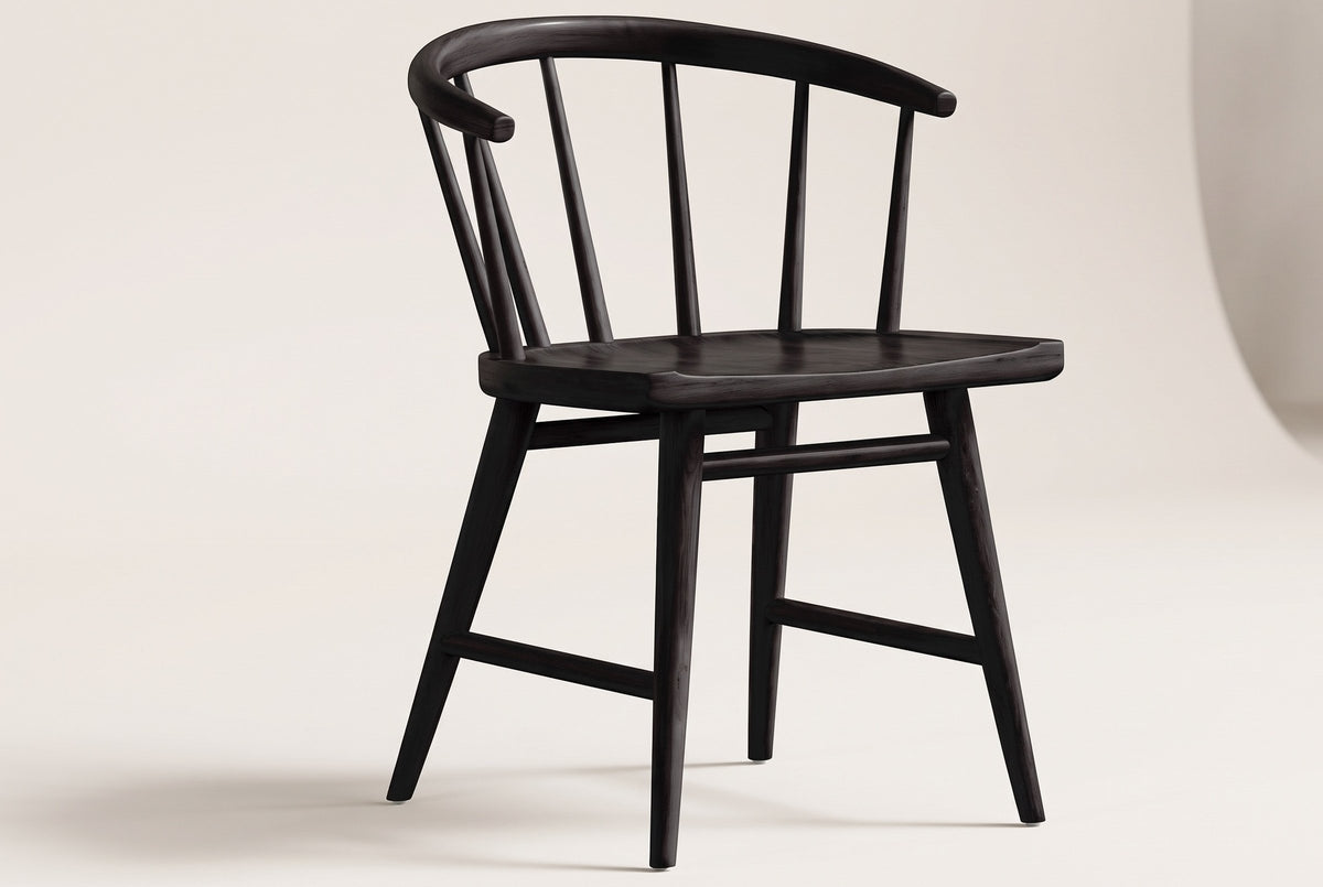 One black oak Lauryn dining chair on an off-white backdrop, seen from the front three-quarter: the curved back rail with rounded tips, the slim spindles, the carved seat and the four splayed legs with their stretchers