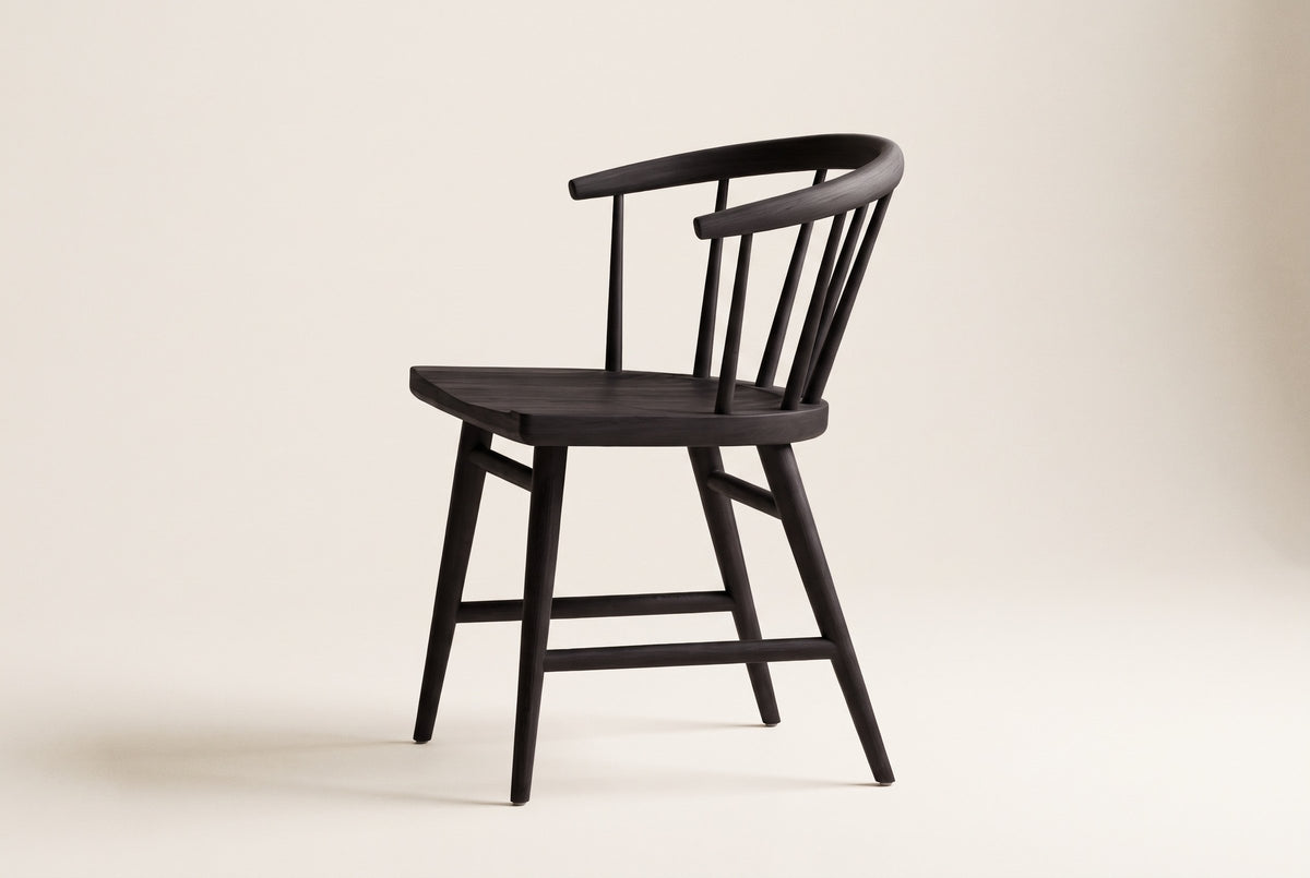Side view of one black oak Lauryn dining chair on an off-white backdrop, with the low curved back rail, the slim spindles, the carved seat and the four splayed legs with their stretchers