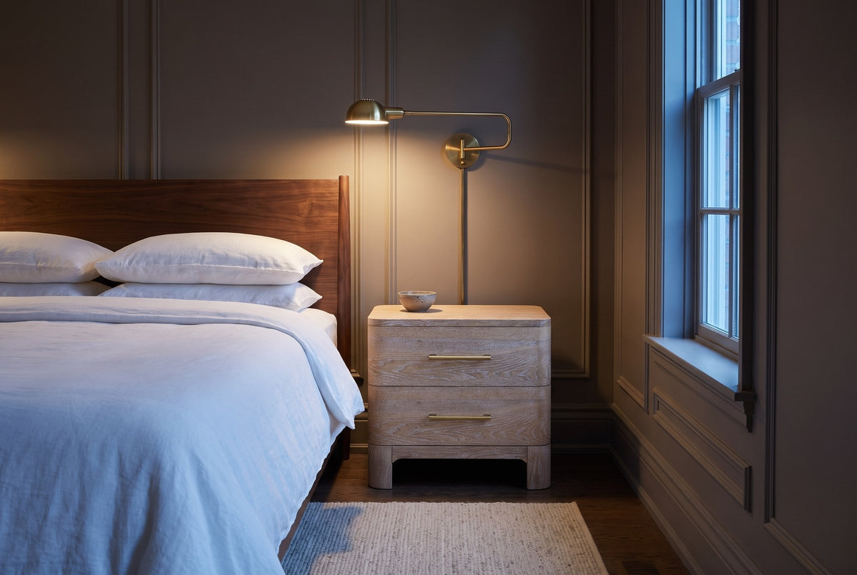 Lodi nightstand in Natural beside a bed in a taupe Ottawa townhouse bedroom at dusk, a brass wall lamp lit above it