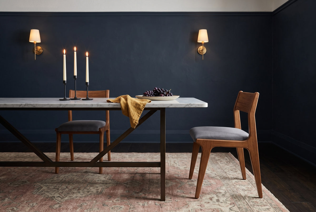 Two Lucrezia dining chairs in the Walnut finish with grey fabric seats at a long white marble table: one tucked in on the far side, the other pulled out past the table end in a side three-quarter view, with three lit taper candles, a mustard linen napkin and a platter of grapes, in a navy room with brass wall sconces and a rose antique rug