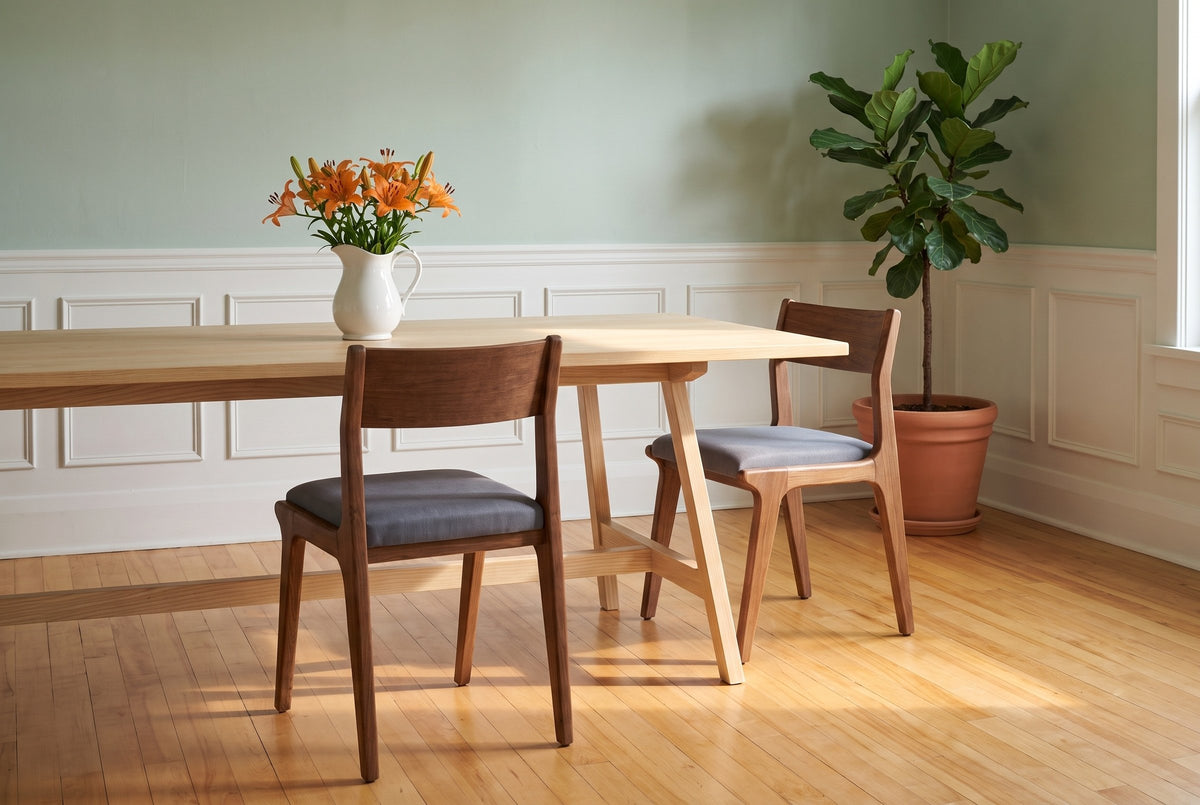 Two Lucrezia dining chairs in the Walnut finish with grey fabric seats at a long pale ash table: one pulled out and seen from behind, the other turned at the far corner of the table in a front three-quarter view, with a white pitcher of orange lilies and a potted fig tree in a celadon green room with white panelled wainscot and honey maple floors in morning sun