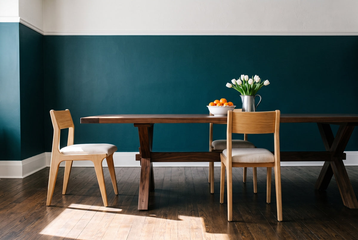 Three Lucrezia dining chairs in the White Oak finish with off-white fabric seats around a long black walnut trestle table: one pulled out at the left end in a side view, one tucked in on the far side and one seen from behind on the near side, with a bowl of clementines and a pewter jug of white tulips, in a deep teal room with dark oak floors in late-morning sun
