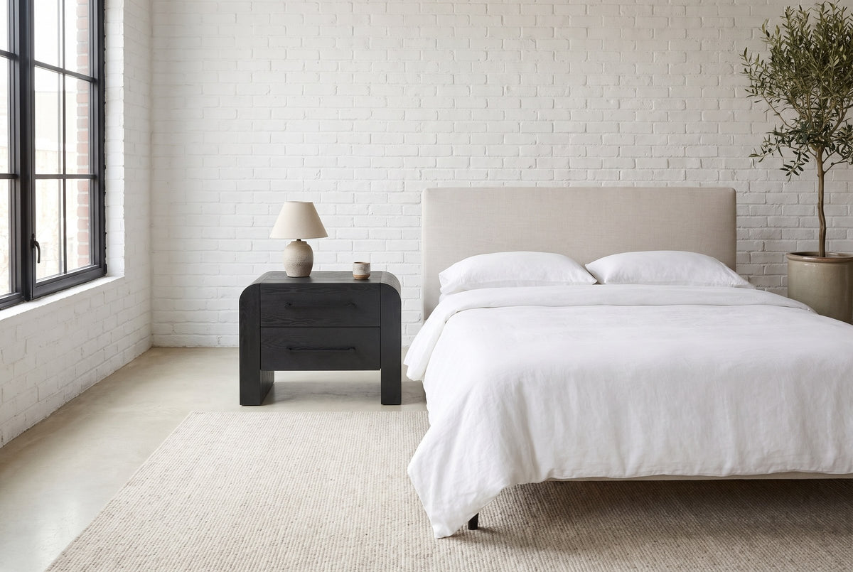 Macerata nightstand in black beside a bed in white linen, in a white-brick loft bedroom with a tall black-framed window