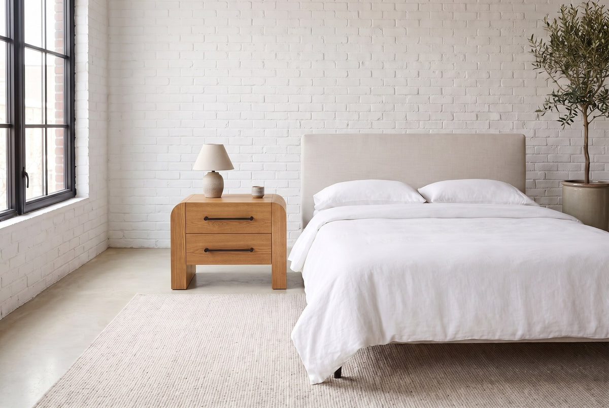 Macerata nightstand in brown beside a bed in white linen, in a white-brick loft bedroom with a tall black-framed window
