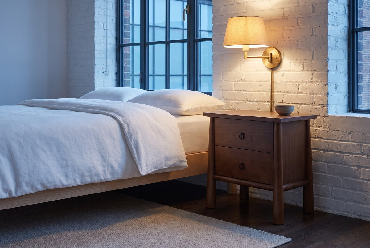 Malcesine nightstand against a whitewashed brick wall in a Montreal loft at dusk, a brass wall lamp lit above it