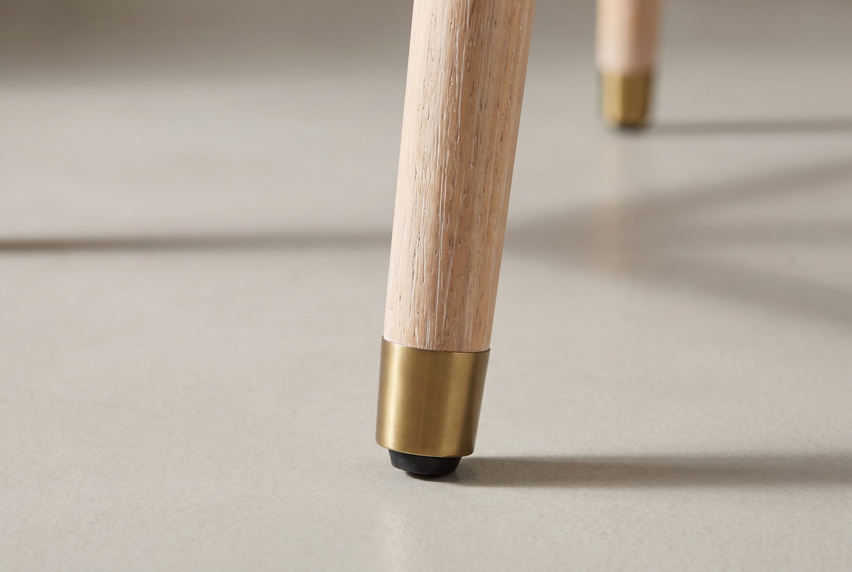 Close-up of one tapered Monteriggioni leg in brushed natural wood ending in a gold-tone foot cap with a black glide