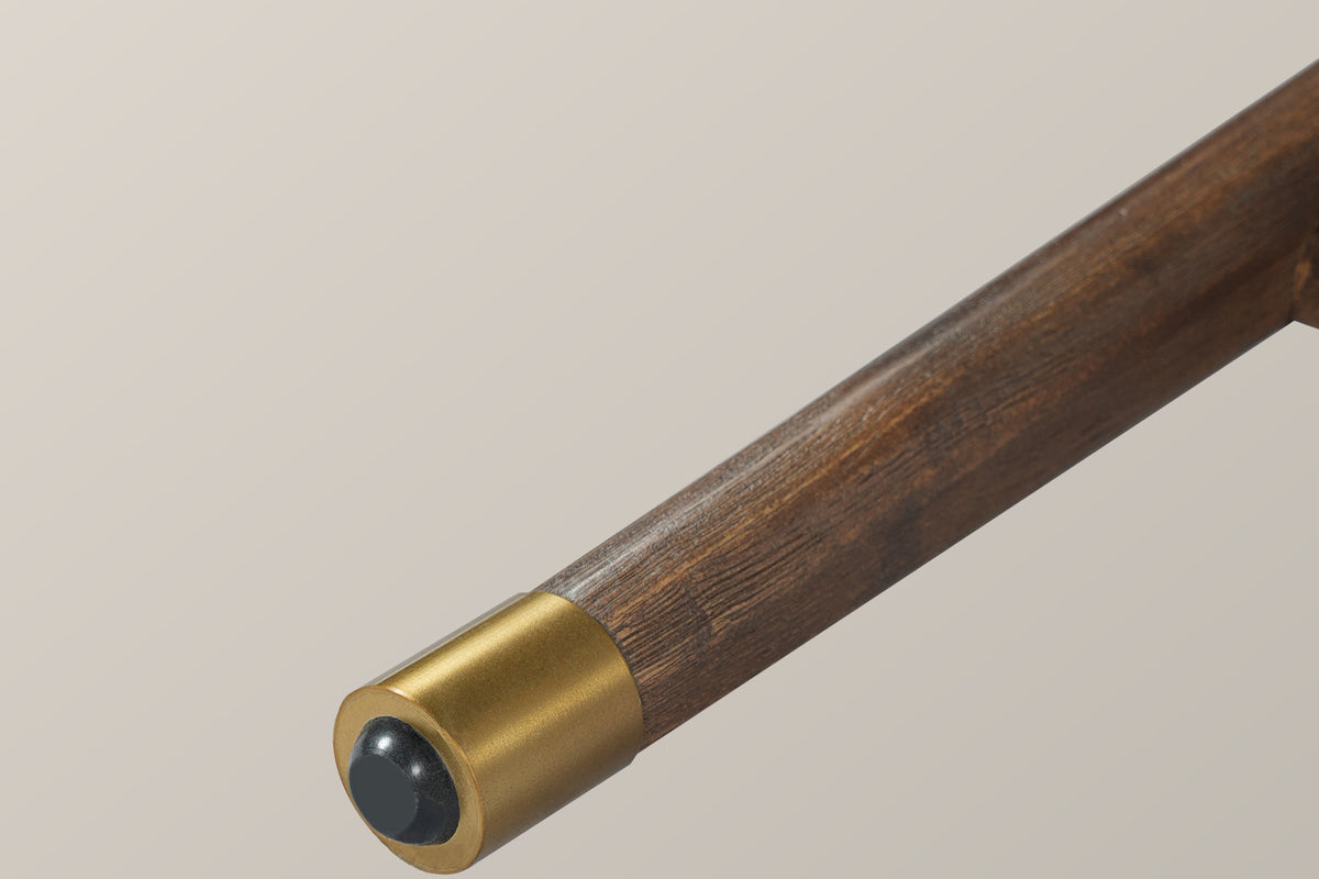 Close-up of one tapered Monteriggioni leg in brushed walnut-finish wood ending in a gold-tone foot cap with a black glide