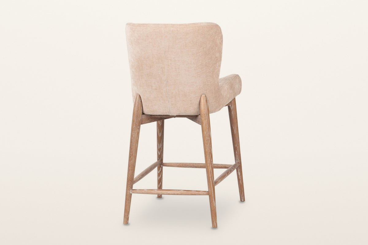 Morrovalle counter stool in Champagne Chenille from behind at an angle on a warm white studio backdrop: the rear legs rise pa