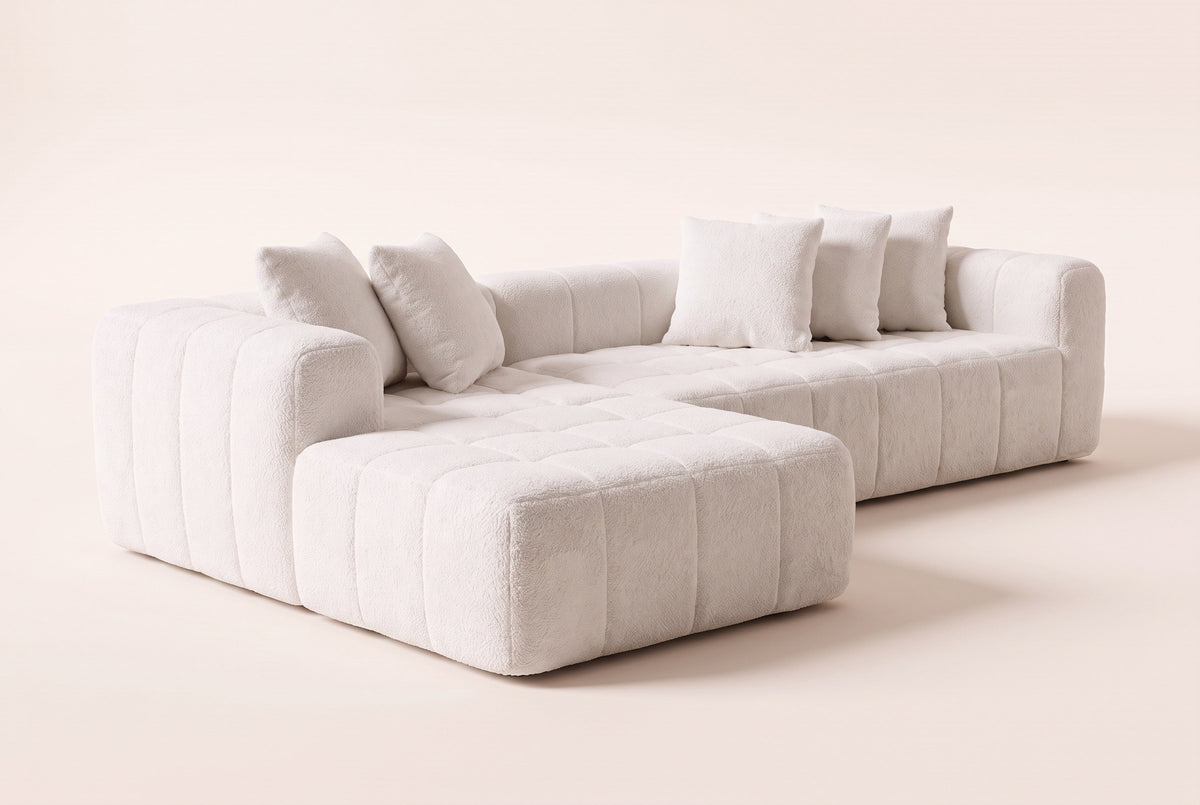 Nembo Boucle Cloud Sectional in white boucle seen from the front-left on a warm off-white backdrop: the chaise nearest, its channelled side and quilted top, the sofa piece behind it