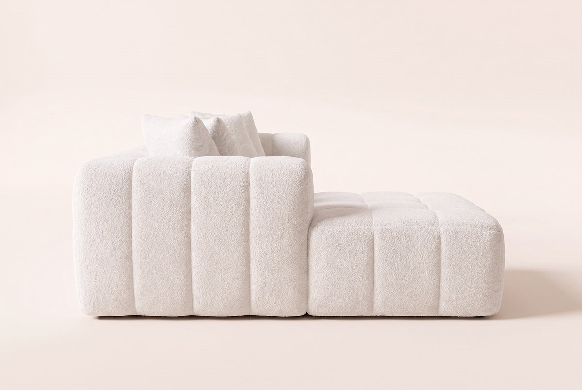 Nembo Boucle Cloud Sectional in white boucle seen from the chaise end on a warm off-white backdrop: the low channelled side sitting straight on the floor, the back and pillows rising behind