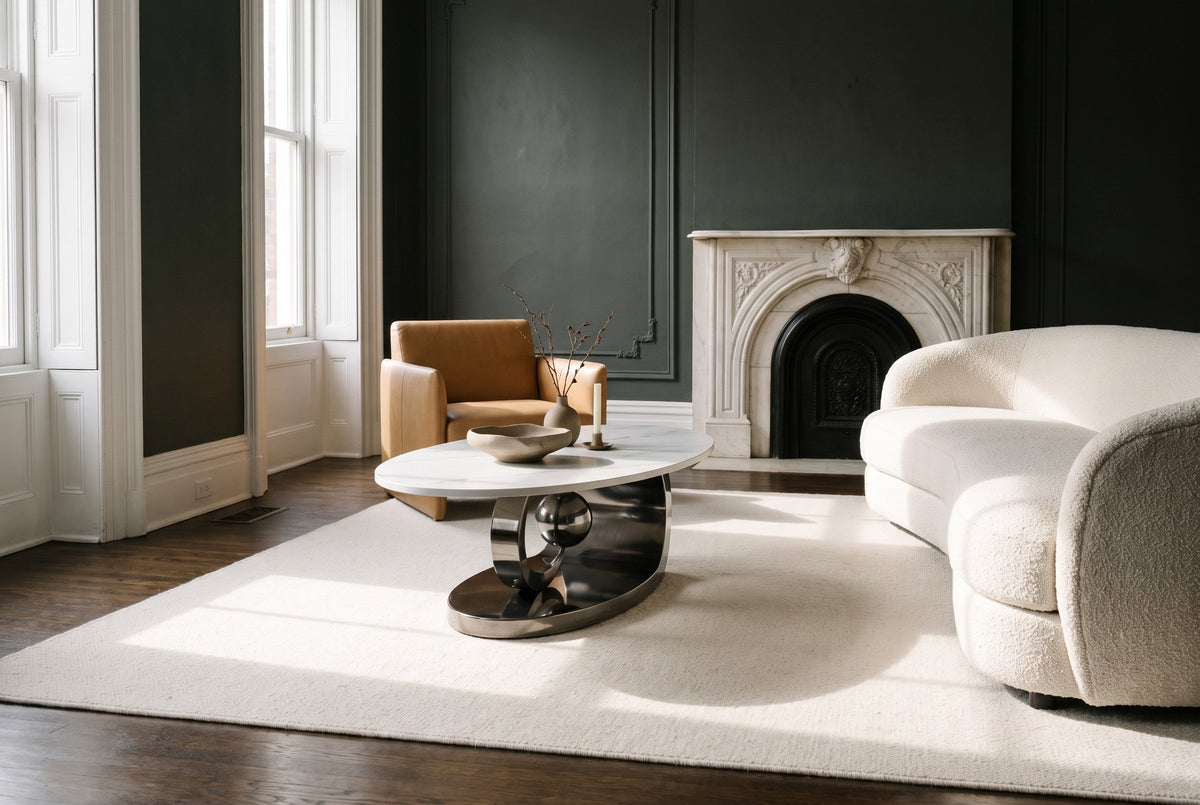 The Oriana coffee table in Gray on an ivory rug in a parlour with charcoal-green plaster walls, a white marble fireplace, an ivory boucle sofa and a camel leather chair