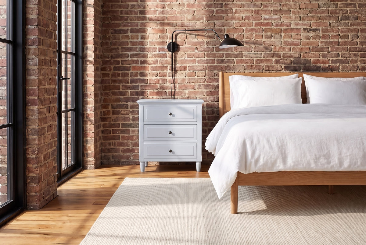 Ortona nightstand in White against an old brick wall in a Montreal loft bedroom, a swing-arm lamp above it