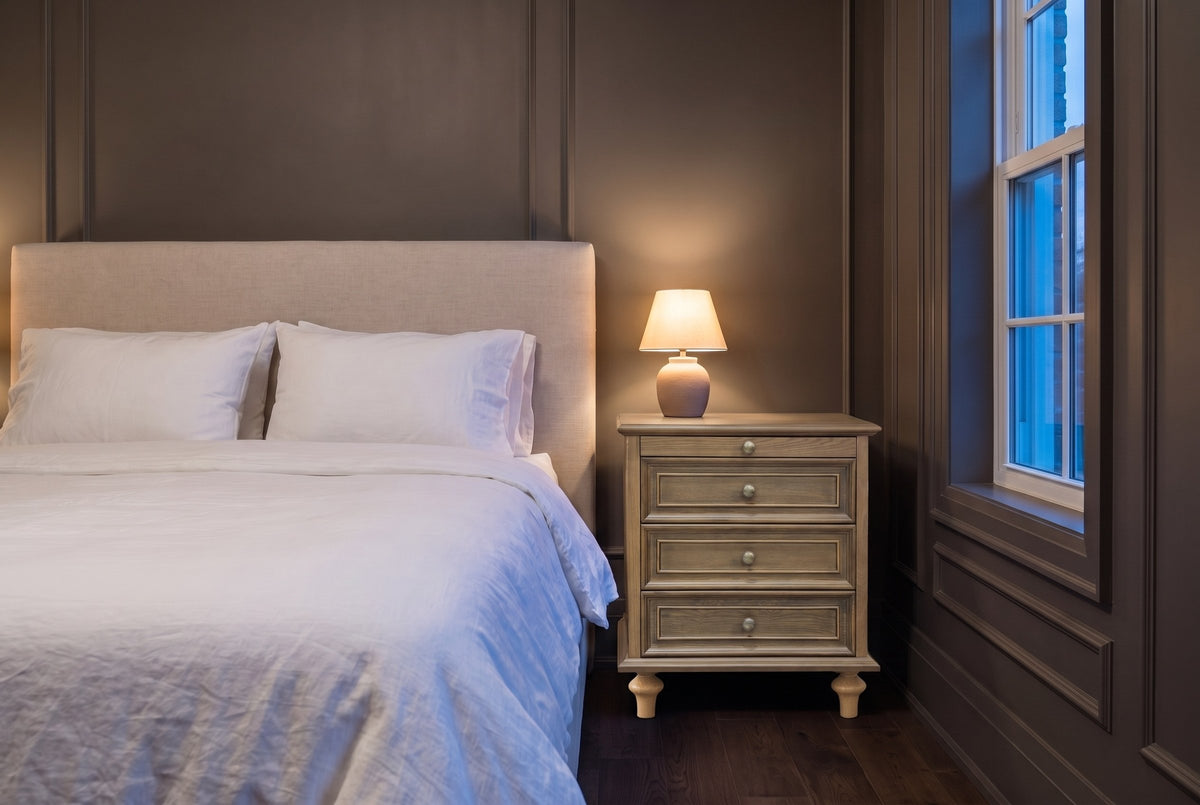 Osimo nightstand in Light Brown in a grey panelled bedroom at dusk, a small lamp lit on top