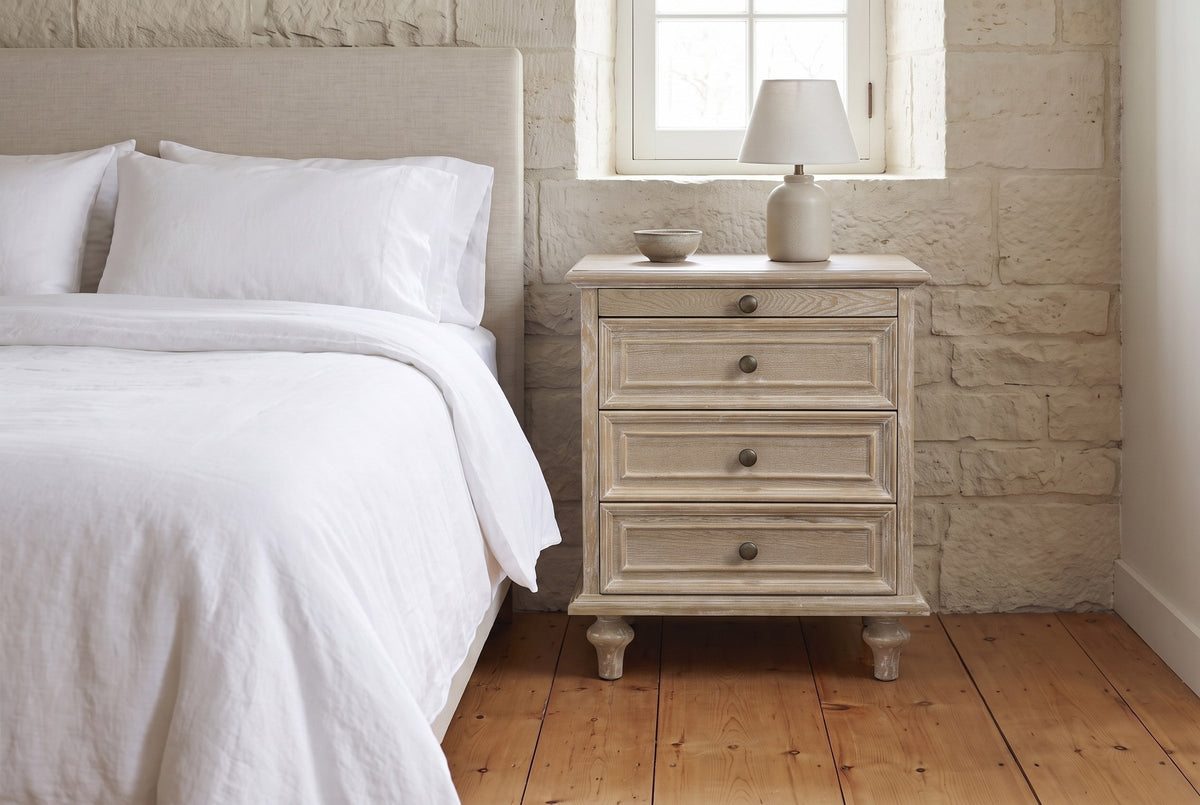 Osimo nightstand in Natural White beside a bed with white linen against a limestone wall, a lamp and a stoneware bowl on top