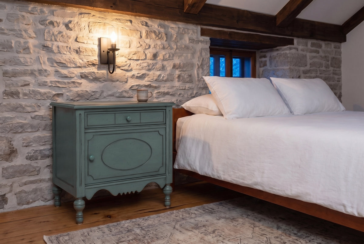 Perugia nightstand in Aged Blue against a fieldstone wall in a Quebec City bedroom at dusk, an iron sconce lit above it