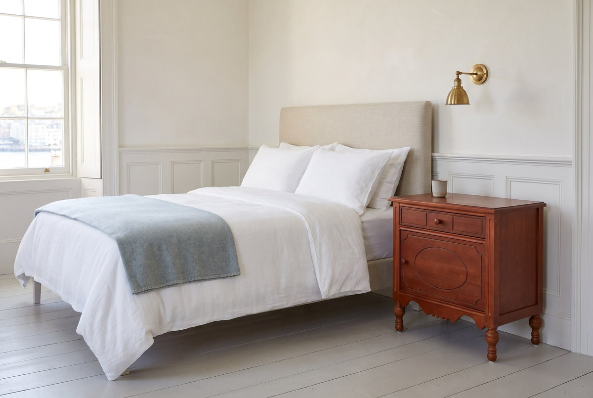 Perugia nightstand beside a bed with white linen in a bright Halifax bedroom with white wainscot and a brass sconce