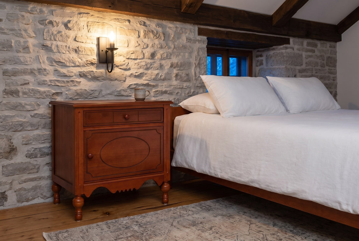 Perugia nightstand against a fieldstone wall in a Quebec City bedroom at dusk, an iron sconce lit above it