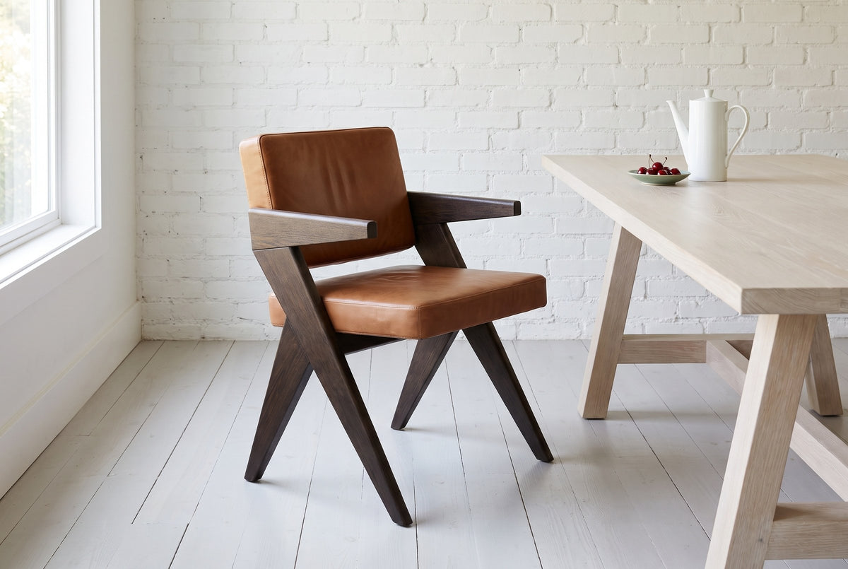 Pierre Jeanneret-style dining chair in dark brown oak and cognac leather beside a pale bleached oak trestle table with a white porcelain coffee pot and a dish of cherries, against a white painted brick wall, on pale painted floorboards by a window in bright daylight