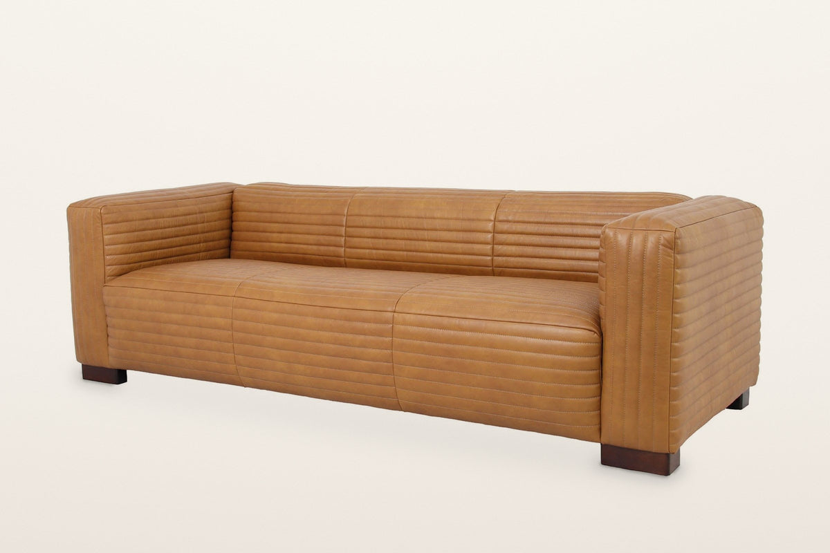 Pontremoli three-seat sofa in Latte top-grain leather with horizontal stitched channels on every face, three-quarter view on a warm white studio backdrop