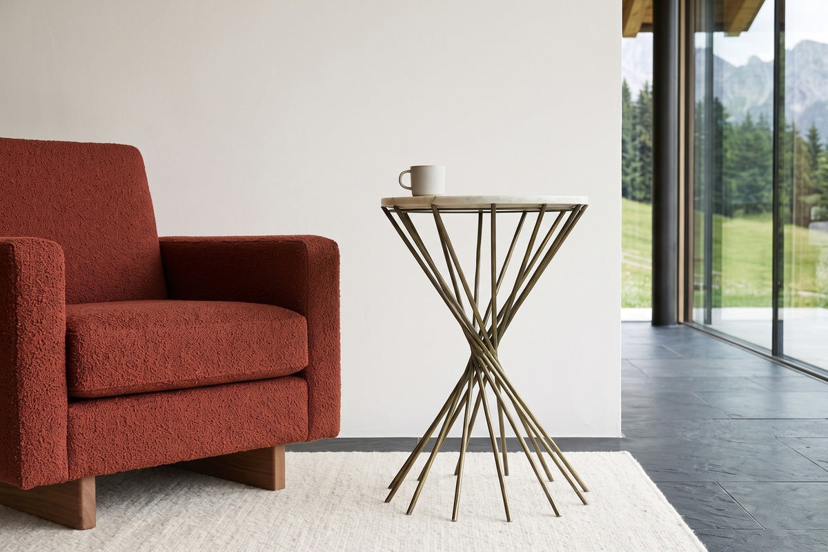 Side view of the Rango side table beside a rust boucle armchair in a Gstaad summer chalet, with white plaster