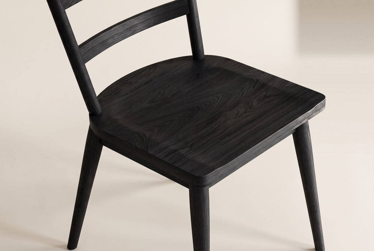 Close-up of one black wire-brushed oak two-slat Rocco dining chair on an off-white backdrop: the sculpted seat and its grain, the lower back slat, the round back posts and the tapered legs