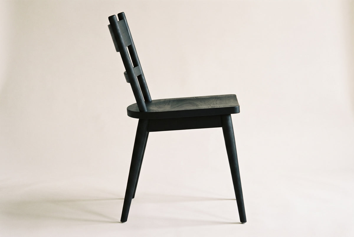 Side view of one black wire-brushed oak two-slat Rocco dining chair on an off-white backdrop, with the back posts leaning back, the sculpted seat and the splayed tapered legs