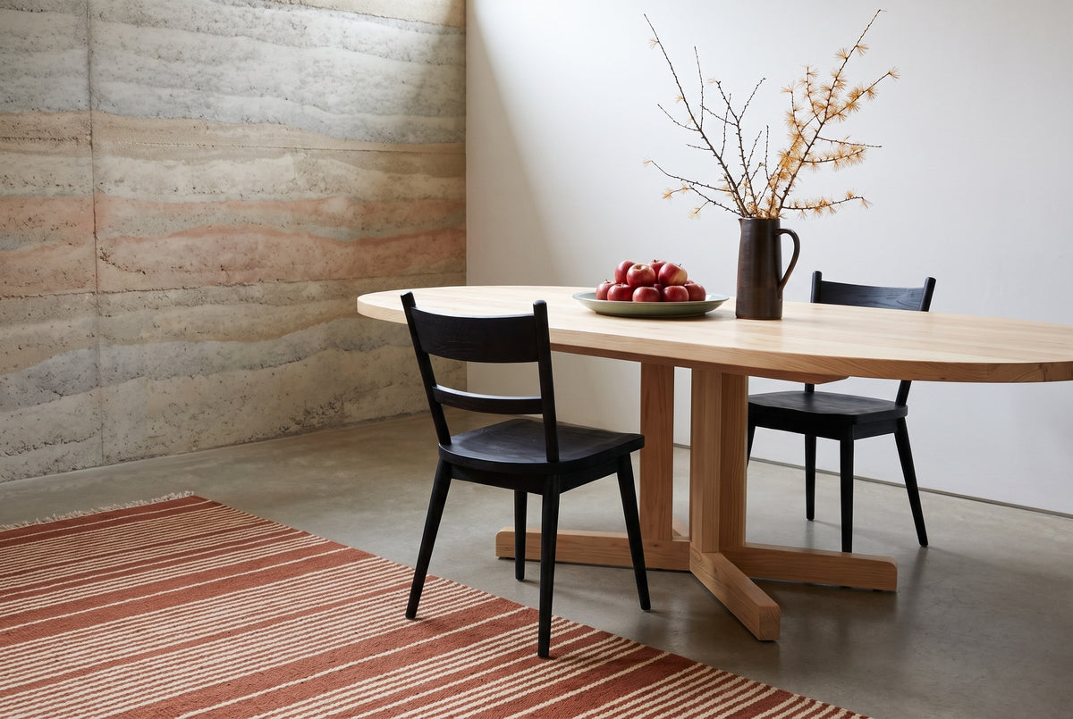 Two black wire-brushed oak two-slat Rocco dining chairs at an oval pale wood table on a cross base, with a platter of red apples and a dark jug of golden larch branches, beside a rammed earth wall on a concrete floor with a rust and cream striped rug