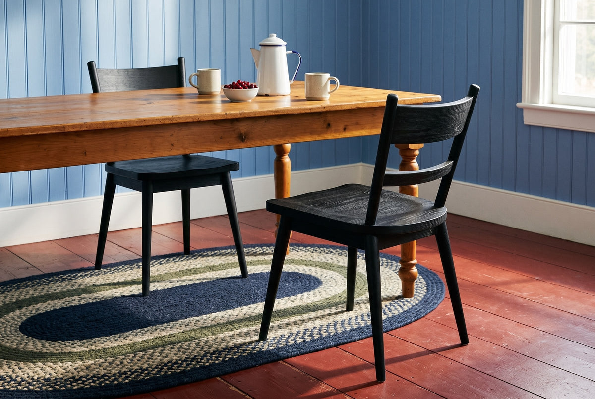 Two black wire-brushed oak two-slat Rocco dining chairs at an antique pine table with turned legs, with a white enamel coffee pot, two cream mugs and a bowl of red berries, in a blue board-walled kitchen with a red painted floor and an oval braided rug in morning sun