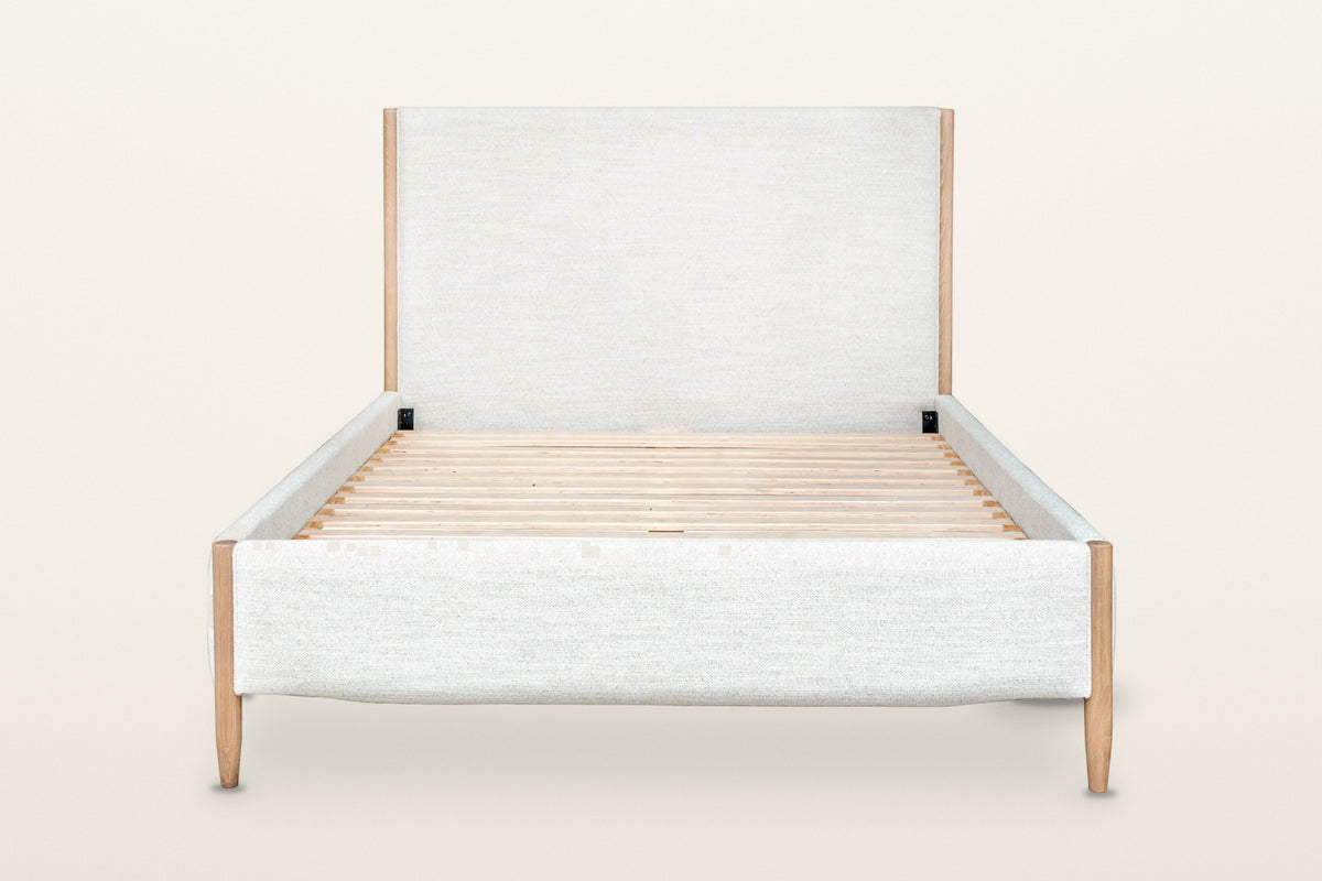 Sarnano bed, front view from the foot on a warm white studio backdrop: the upholstered foot rail between two round oak posts