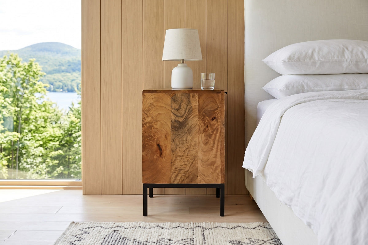 Side view of the Sarteano nightstand in Pecan mango next to a bed in an Orford lake house bedroom