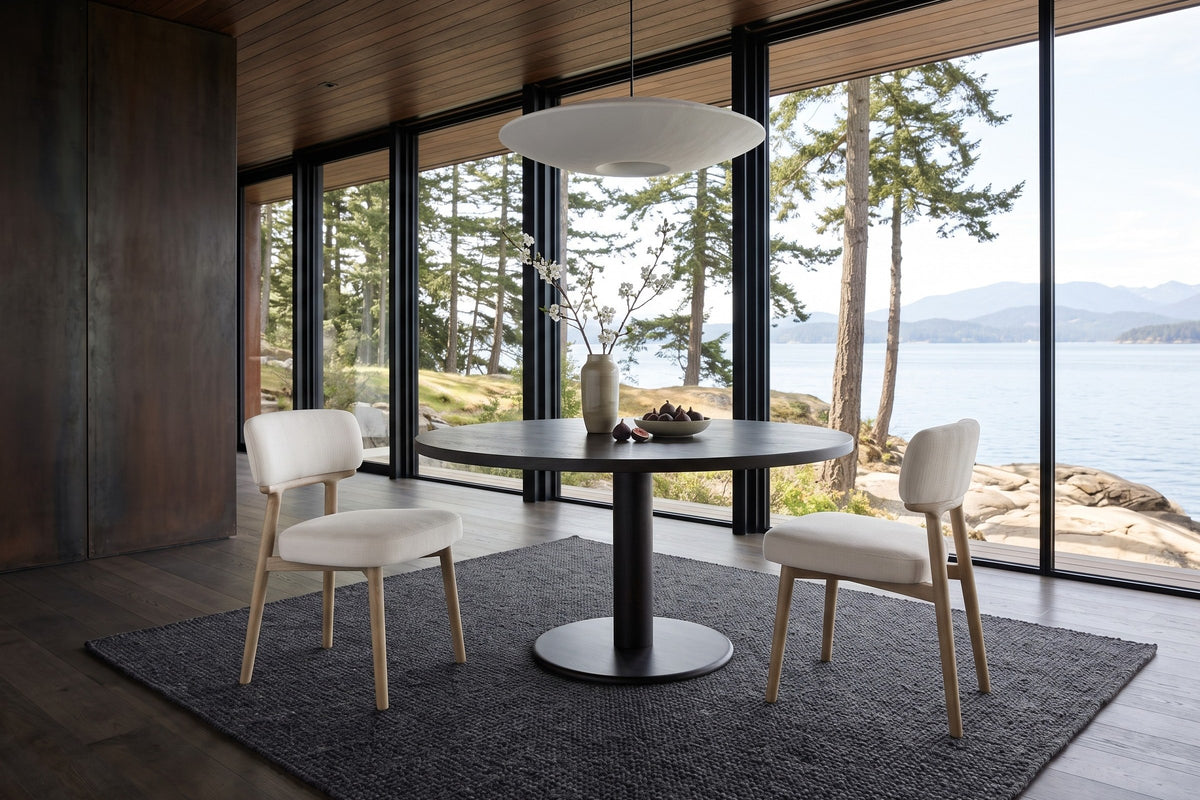 Two Savoca dining chairs in Porcelain Chenille at a round dark oak table in a Galiano Island house, with a dark corten wall