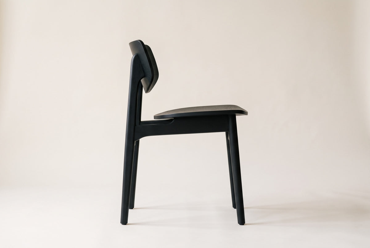 Black Simone dining chair in side view on a plain warm off-white studio backdrop, showing the curved back panel raised on the rear posts, the open gap above the seat, the side rail and four slim legs