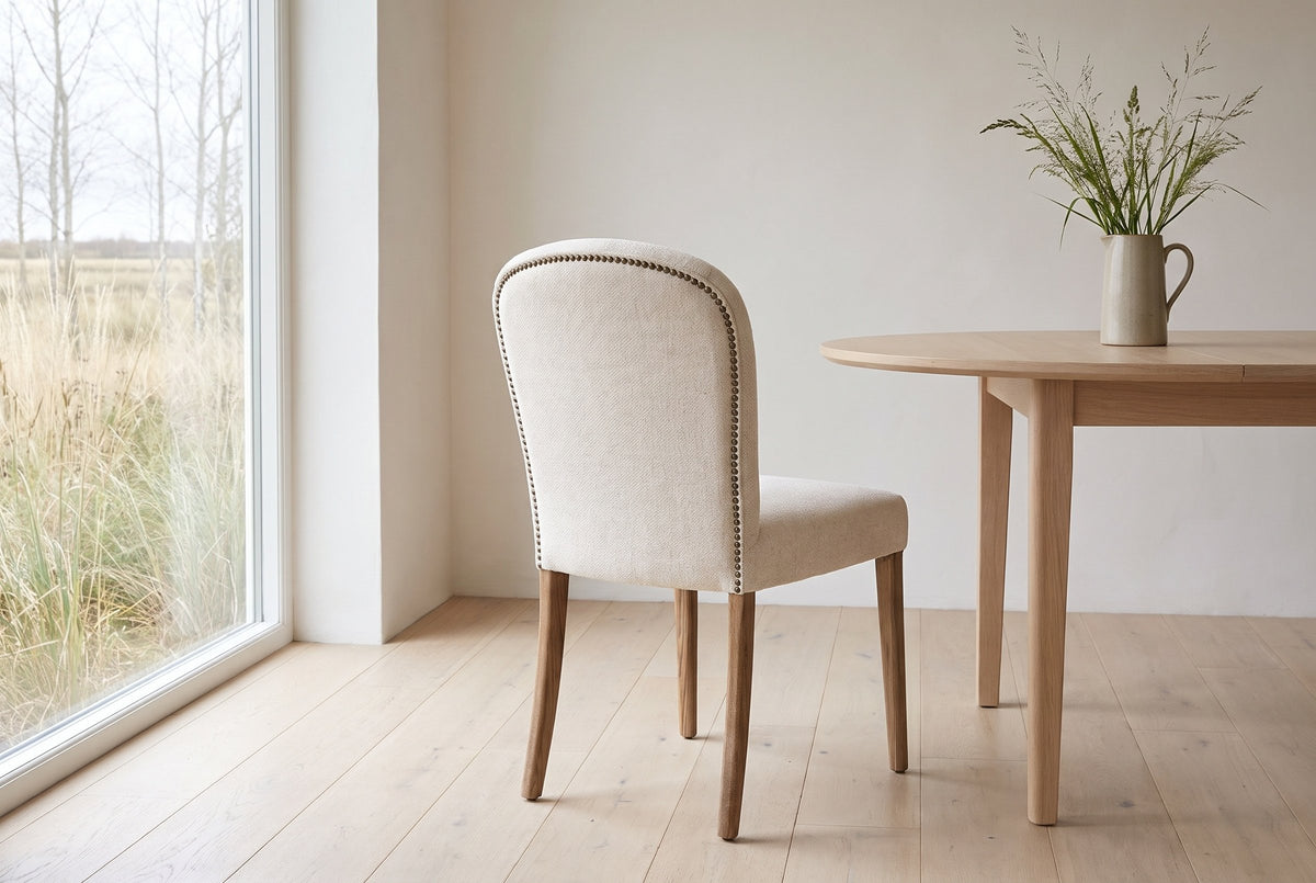 Usseglio dining chair seen from behind, the row of antique-bronze nailheads round its cream back, beside a pale oak round table in a Saskatoon dining room