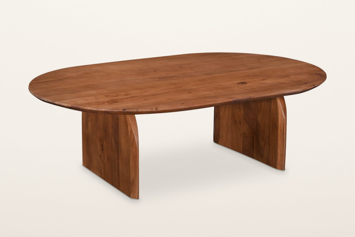 Visso solid acacia coffee table in Pecan, three-quarter view on a warm white studio backdrop: a soft oval top on two arched p