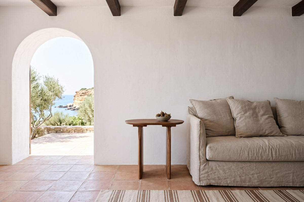 Side view of the Visso side table in Pecan at the arm of a linen sofa in a stone house by the sea, with white lime plaster