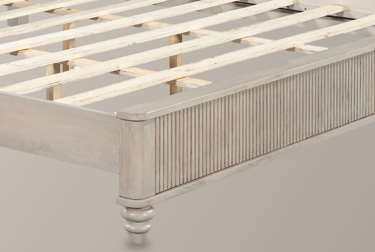 Close-up of the beige reeded footboard corner with its rounded cap and a turned bun foot, the pale slats behind