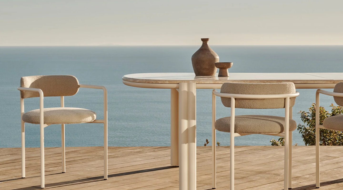 This image depicts a serene outdoor setting by the sea, featuring a round patio table and matching chairs. The table has a sleek, light - colored surface and modern legs, while the chairs have a contemporary design with light - colored frames and cushioned seats and backs. On the table, there are a couple of rustic - looking vases, adding a touch of charm. These pieces are part of the Valencia patio furniture collection, including Valencia patio table chairs and Valencia patio chairs. They are perfect examples of stylish outdoor furniture chairs, combining comfort and aesthetic appeal for an ideal outdoor relaxation experience.