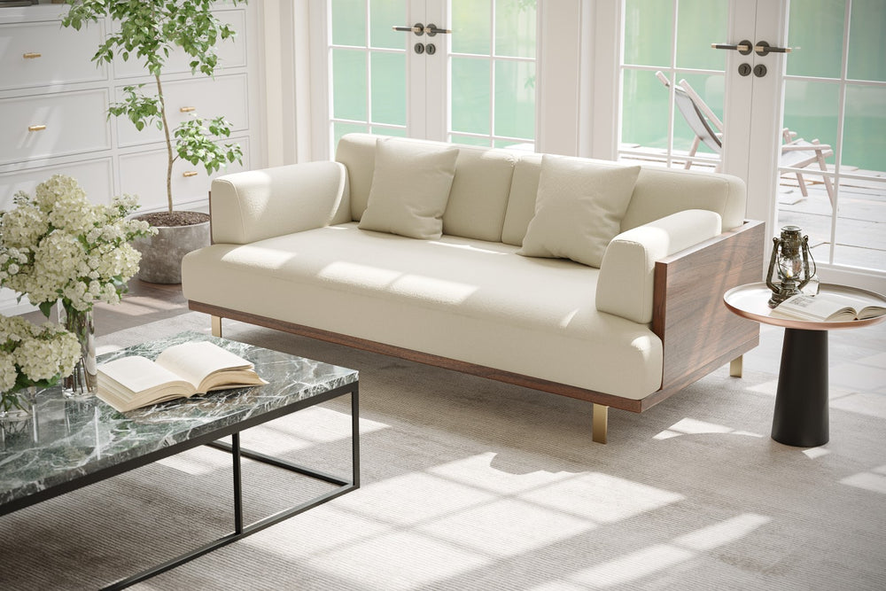 In a Living Room, There is Right Acute Angle Front View of A Luxurious, Beige, Kiln Dried Wood Frame, Emilia Modern Fabric Sofa with Two Cushions on It, and Also There is a  Plant in a pot in the Sofa Left-Side.