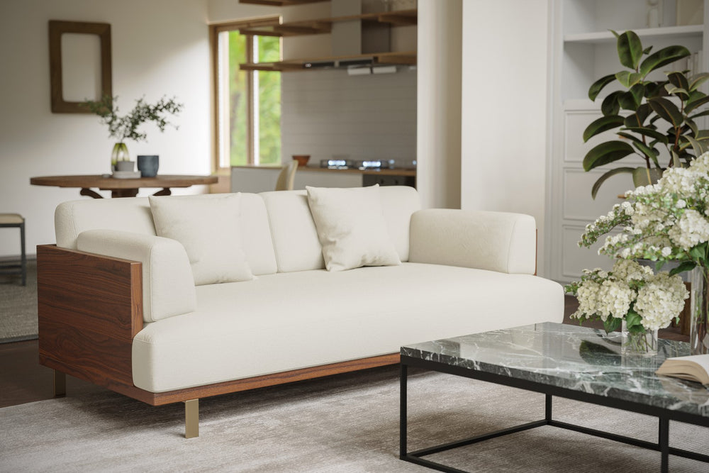 In a Living Room, There is Left Acute Angle Front View of A Luxurious, Beige, Kiln Dried Wood Frame, Emilia Modern Fabric Sofa with Two Cushions on It and Also There is Plant In Glass Pot and Plastic Pot.